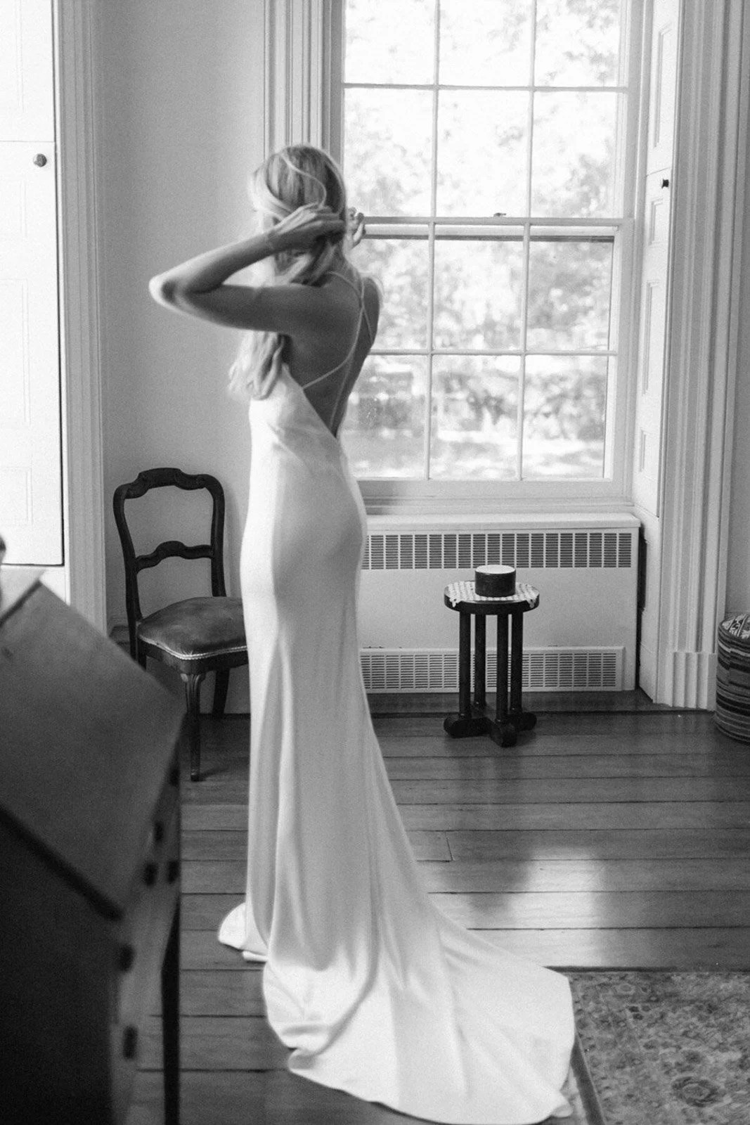 A bride in a long, elegant white dress standing near a large window, adjusting her hair.