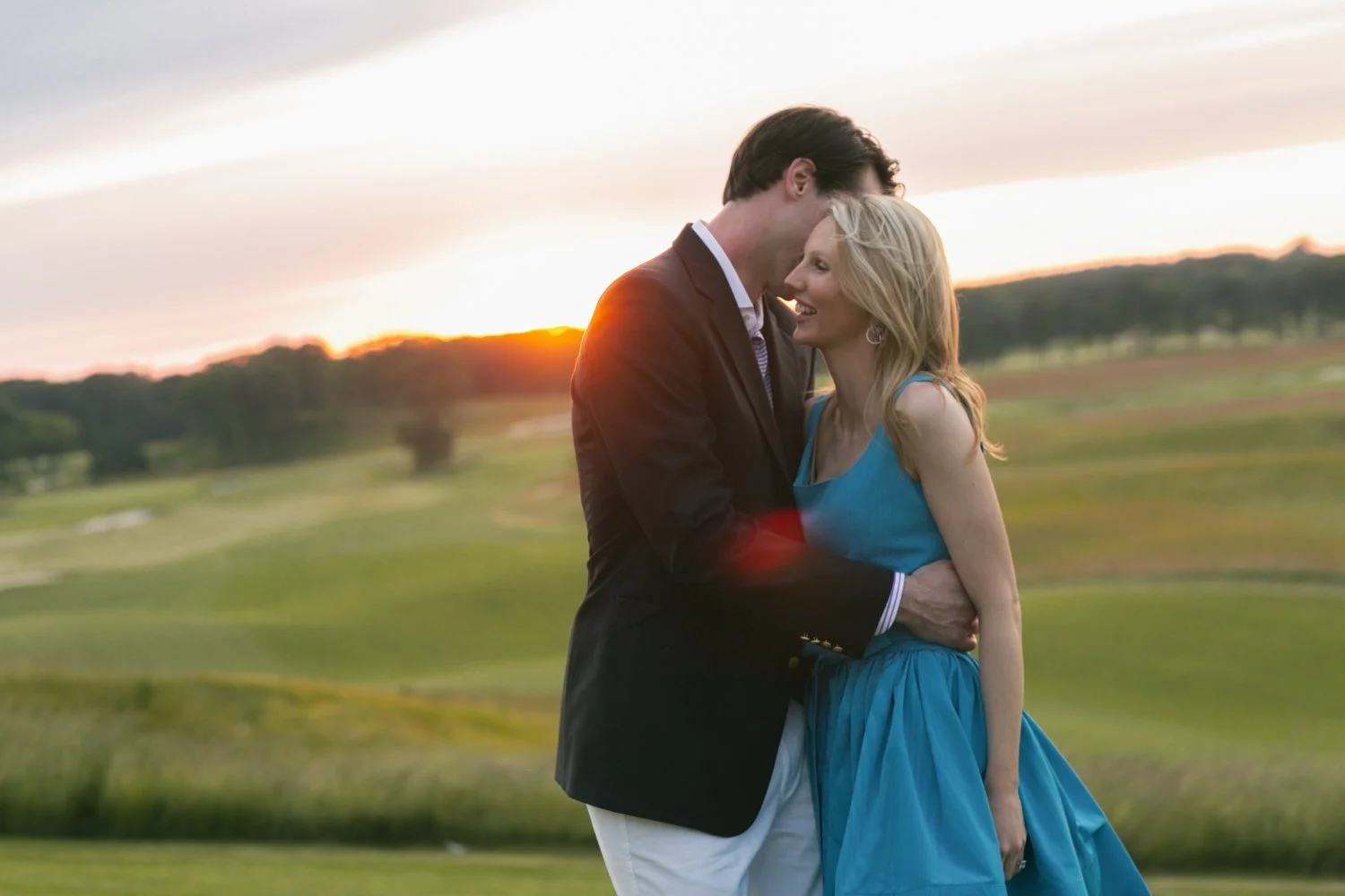 A man and woman embrace and smile in a field at sunset, the woman wearing a blue dress and the man in a dark jacket, with a scenic landscape in the background.