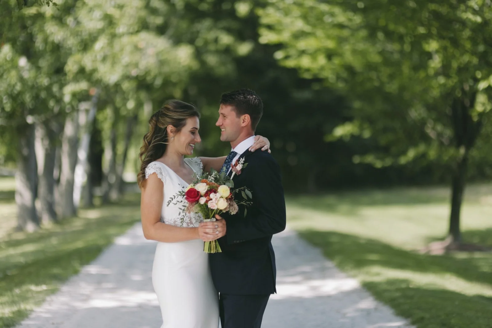 A newlywed couple is smiling and looking into each other's eyes while standing on a tree-lined path outdoors. The bride is holding a bouquet of flowers, and they are embracing each other.