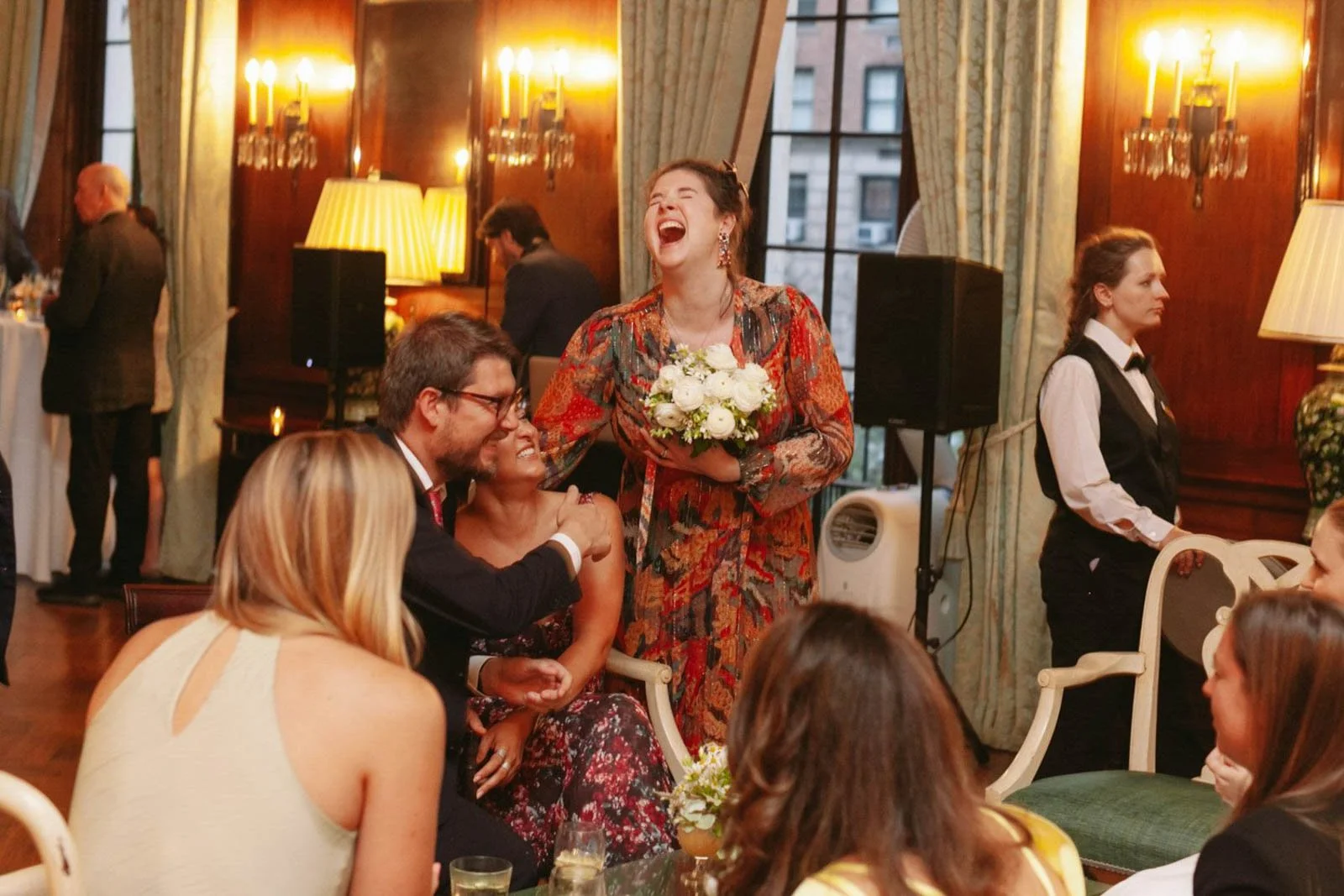 A woman holding a bouquet of white flowers laughing joyfully while people sit around her in a warmly lit elegant room, suggesting a celebration such as a wedding.
