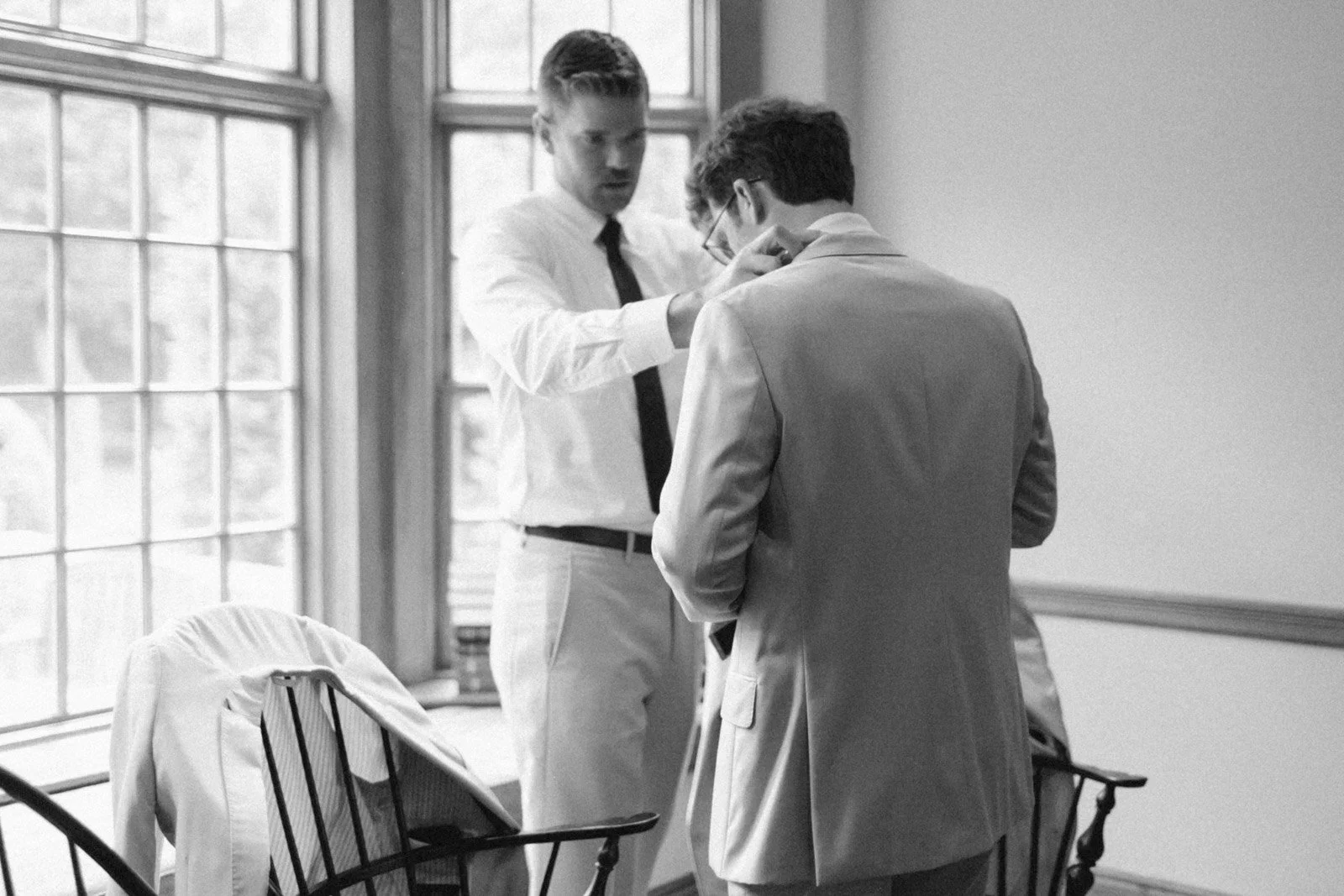 Two men in professional clothing standing in an office with large windows. One man is adjusting the tie of the other man.