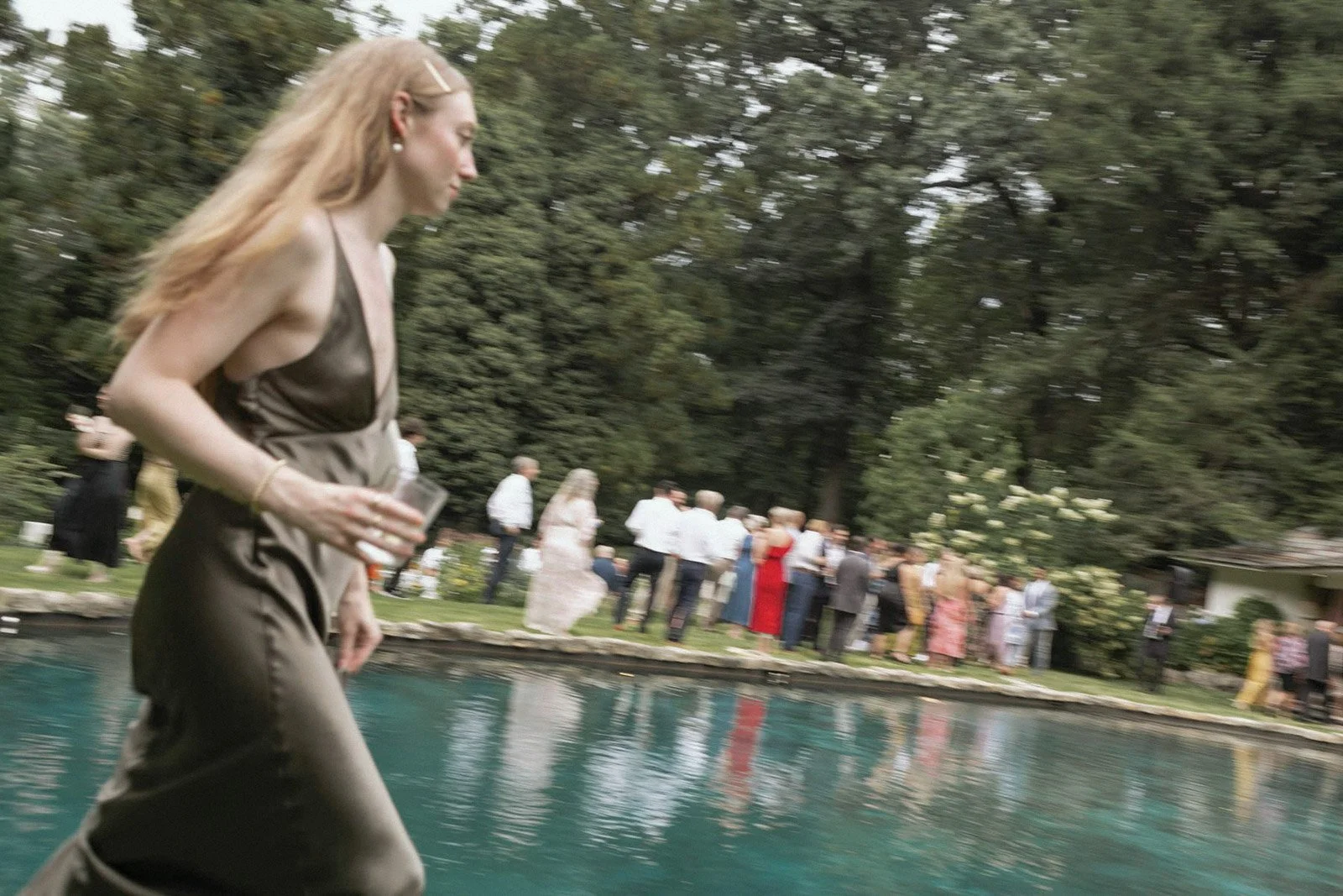 A woman in a brown satin dress is walking near a swimming pool, holding a glass, with a group of people dressed in formal attire in the background, outdoors with trees.
