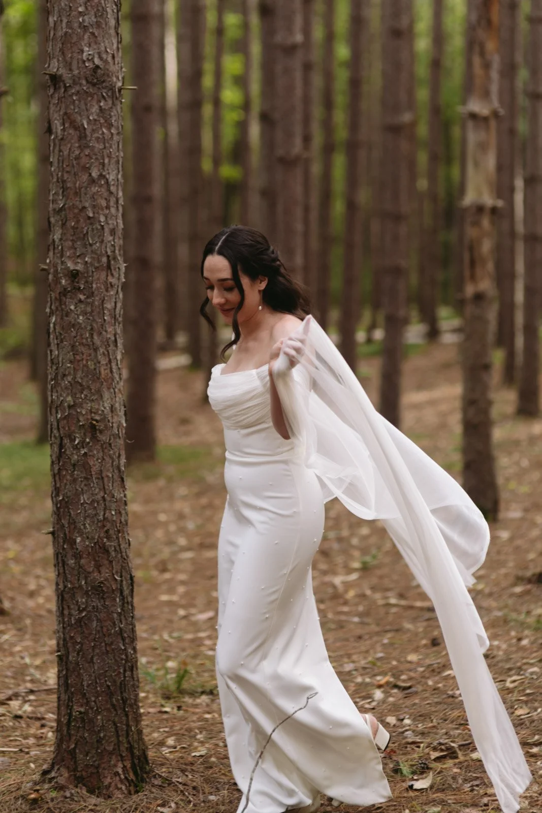 A woman in a white wedding dress walking through a forest of tall trees.