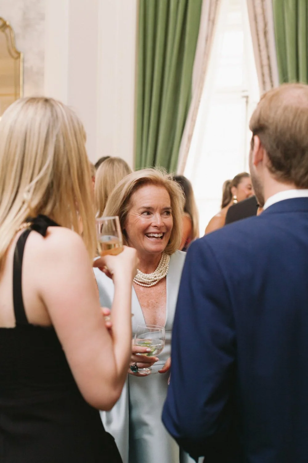 Women at a social event talking and smiling, holding drinks, with green curtains in the background.