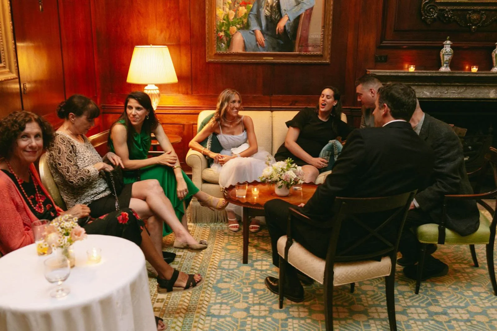 Group of women and men sitting in a cozy, wood-paneled room, engaging in conversation, with a table of flowers and candles, and a portrait hanging on the wall.