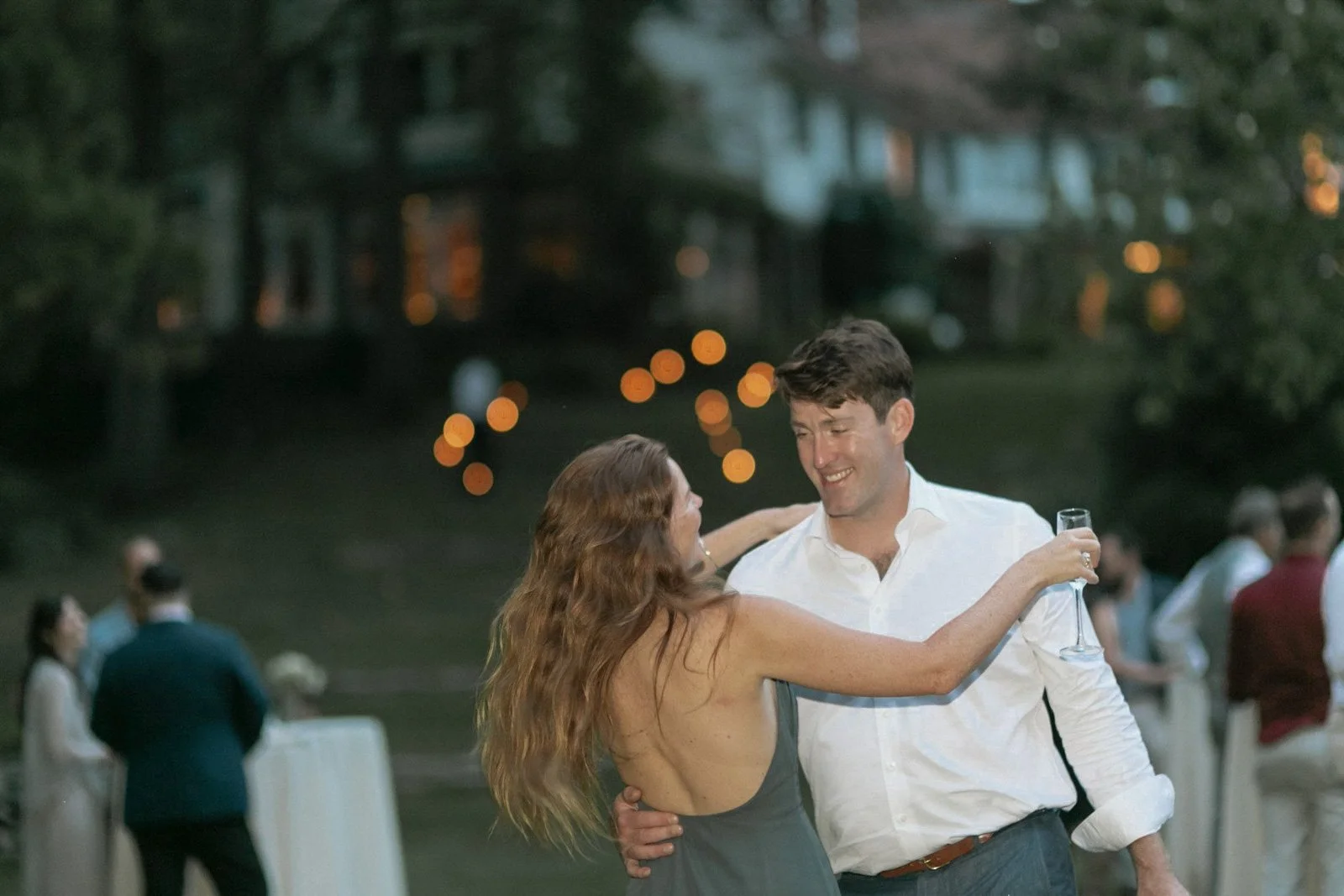 A couple dancing and smiling at an outdoor event, with blurred guests and string lights in the background.