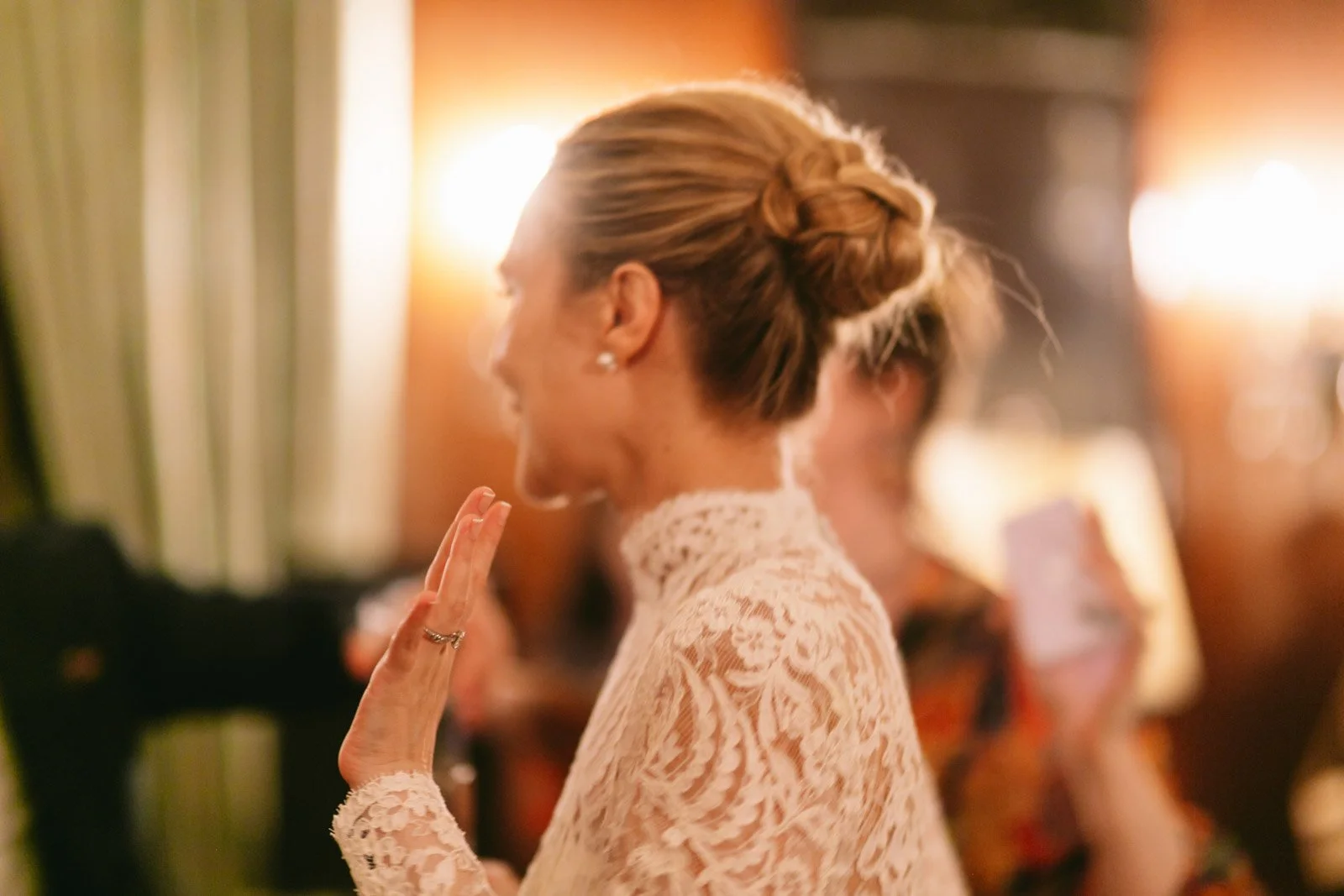 A woman with styled hair in an elegant updo and wearing lace clothing, smiling and waving at an indoor social gathering with warm lighting.