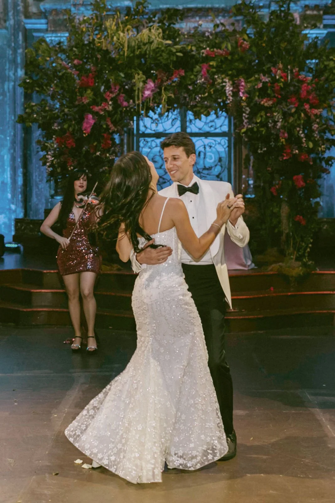 A bride and groom are dancing at their wedding reception. The bride is wearing a white, sequined wedding dress, and the groom is dressed in a tuxedo with a black bowtie. A musician playing a violin is visible in the background, with a floral arch beh