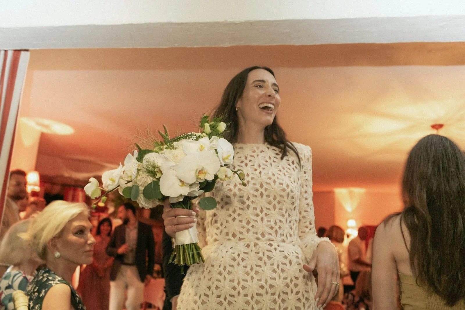 Woman holding a bouquet of white flowers, smiling at a gathering or celebration indoors with warm lighting.