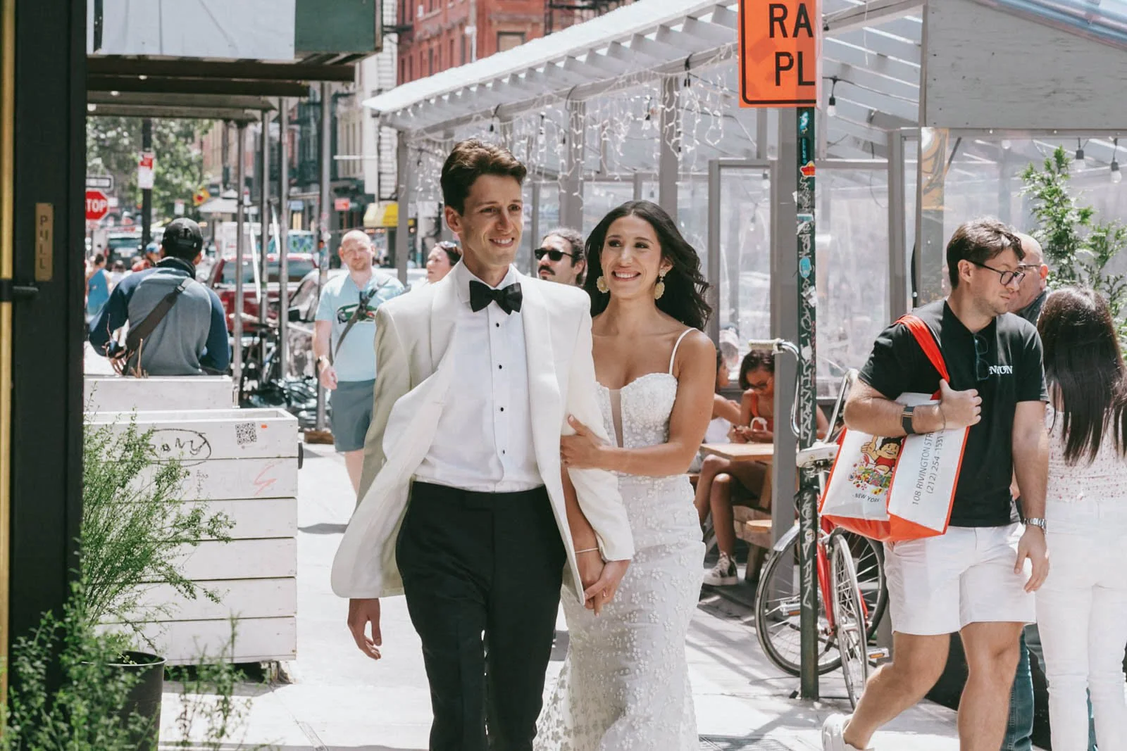 A couple dressed in wedding attire walking on a city sidewalk, surrounded by pedestrians and outdoor seating at a café or restaurant.