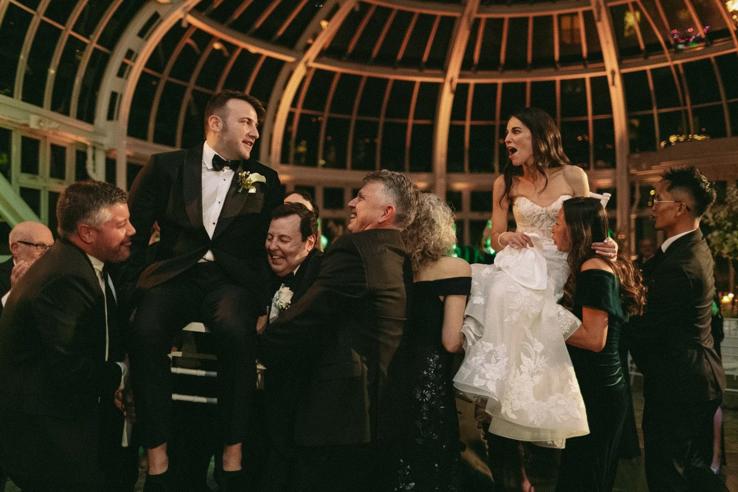 A wedding reception with guests celebrating inside a glass-domed structure, featuring a bride in a white wedding gown and a groom in a black tuxedo, surrounded by friends and family.