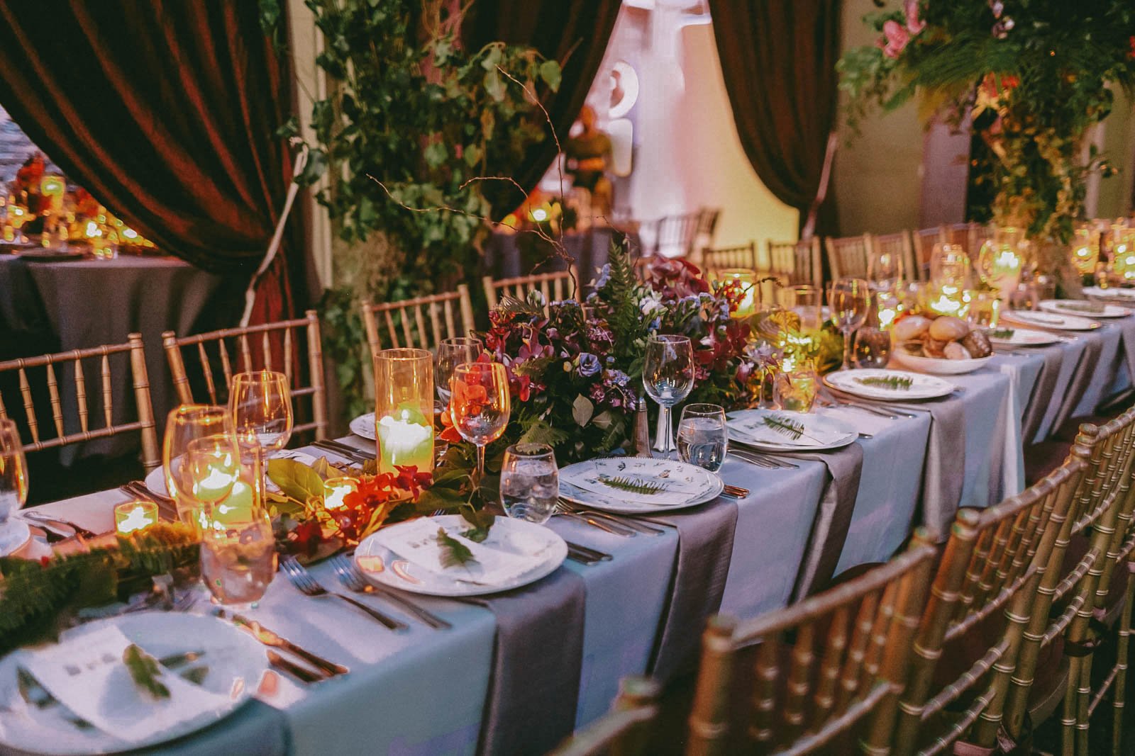 A beautifully decorated banquet table set for an event with floral centerpieces, candles, and elegant tableware, featuring wooden chairs and dark curtains in the background.