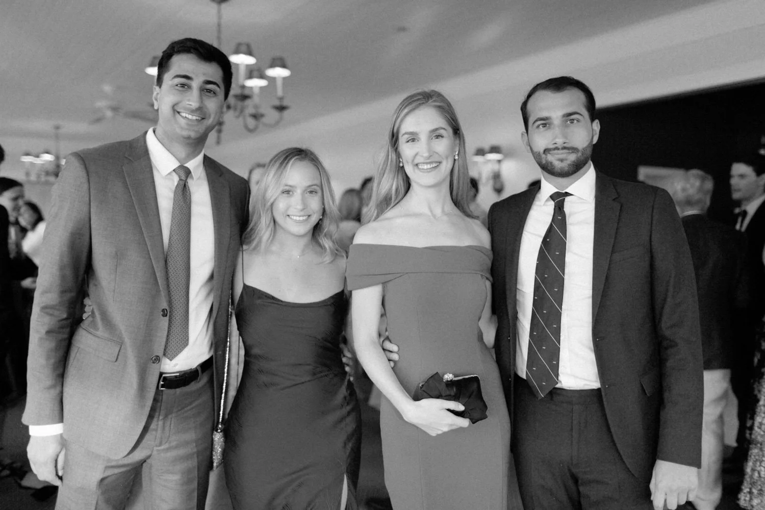 Four people dressed in formal attire, smiling at a social event or party, with a blurred crowd and ceiling lights in the background.