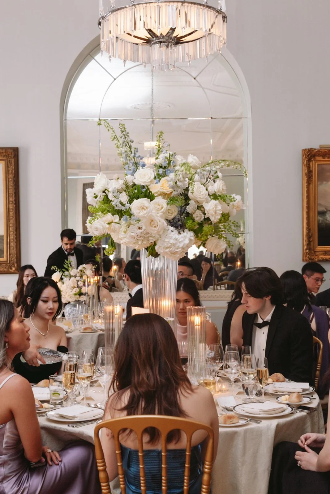 A formal dinner event with guests seated around a table decorated with a large floral centerpiece and candles, in a room with a chandelier and a large mirror.