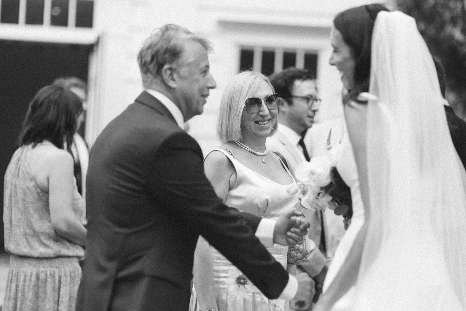 A black-and-white photo of a wedding ceremony with a groom and bride facing each other, with two people in the background. The groom is smiling and shaking hands with the bride, who is wearing a veil and holding a bouquet. The woman in the background