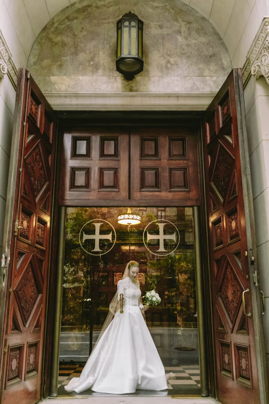 A bride in a white wedding gown holding a bouquet of flowers, standing inside an arched doorway with ornate wooden doors, with a glass window behind her reflecting trees and a building at Christ Church NYC.