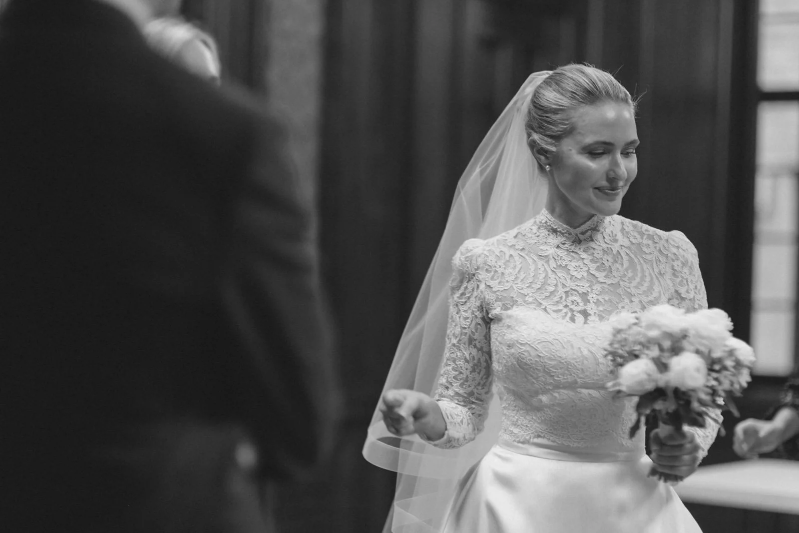 A bride in a lace wedding dress holding a bouquet of flowers, smiling with eyes closed during a wedding ceremony.
