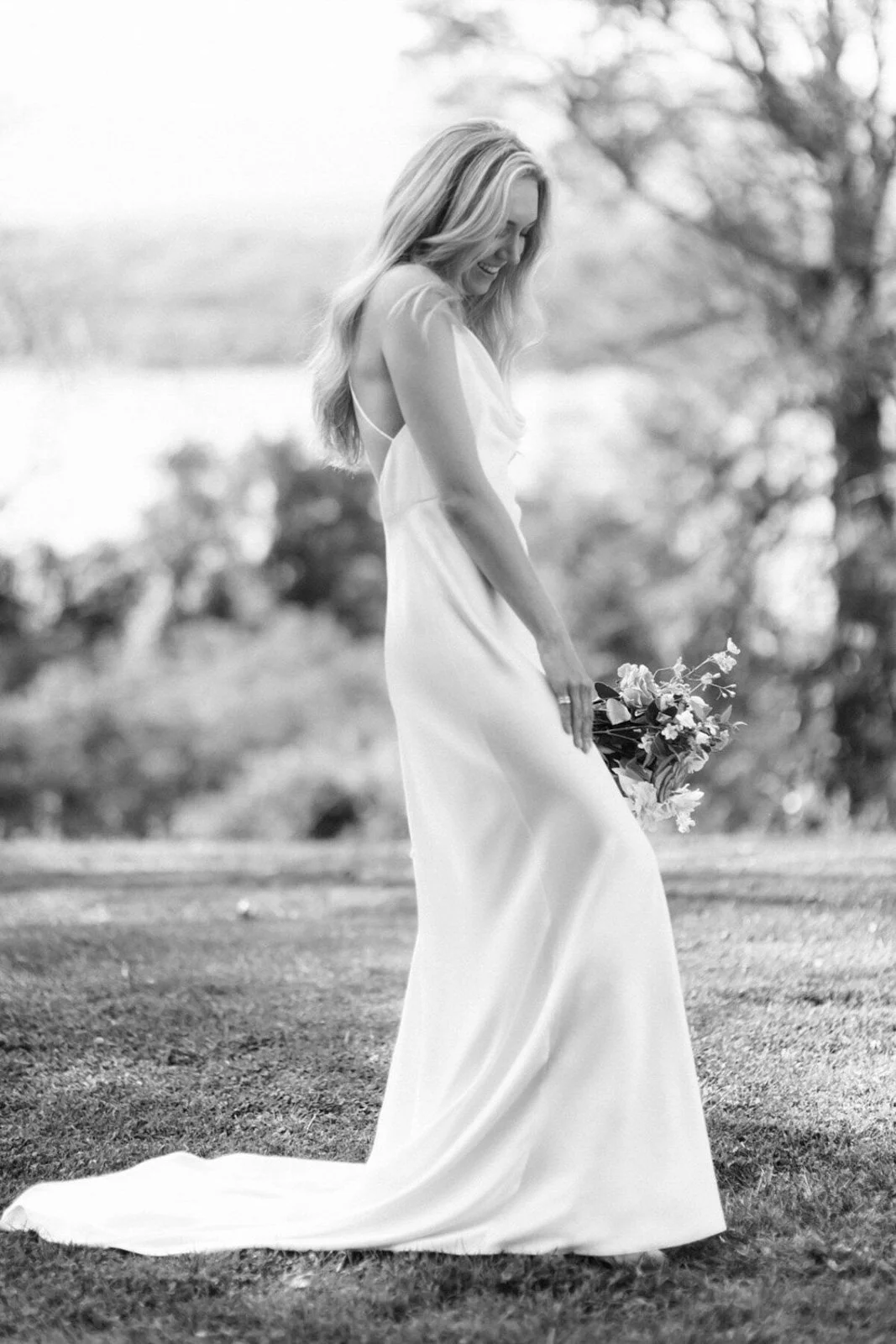 A woman in a long, flowing dress holding a bouquet of flowers outdoors, smiling and looking down, with trees in the blurry background.