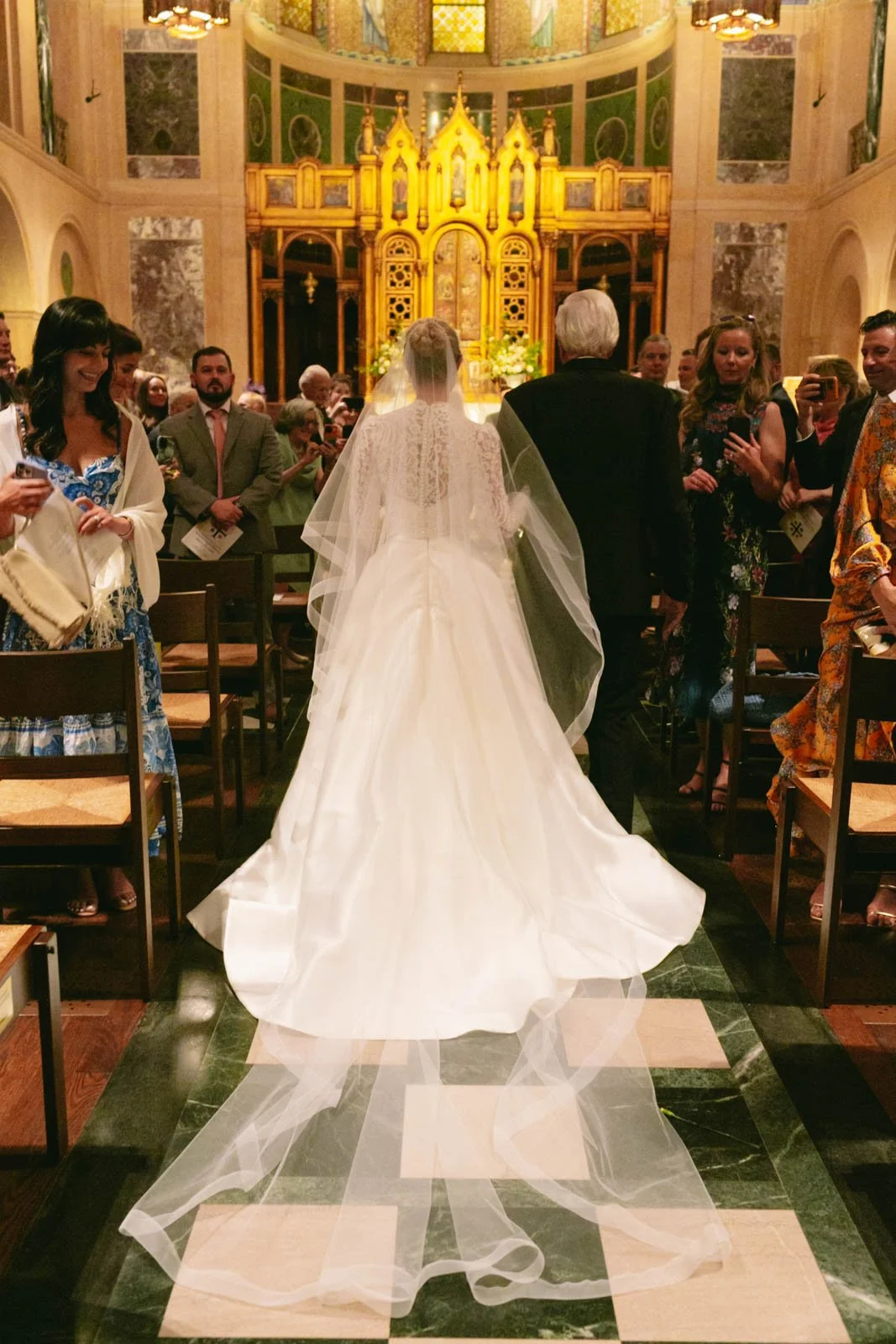 Bride in a white wedding gown and veil walking down the aisle of a church, surrounded by seated and standing guests, with an ornate altar backdrop in the background.