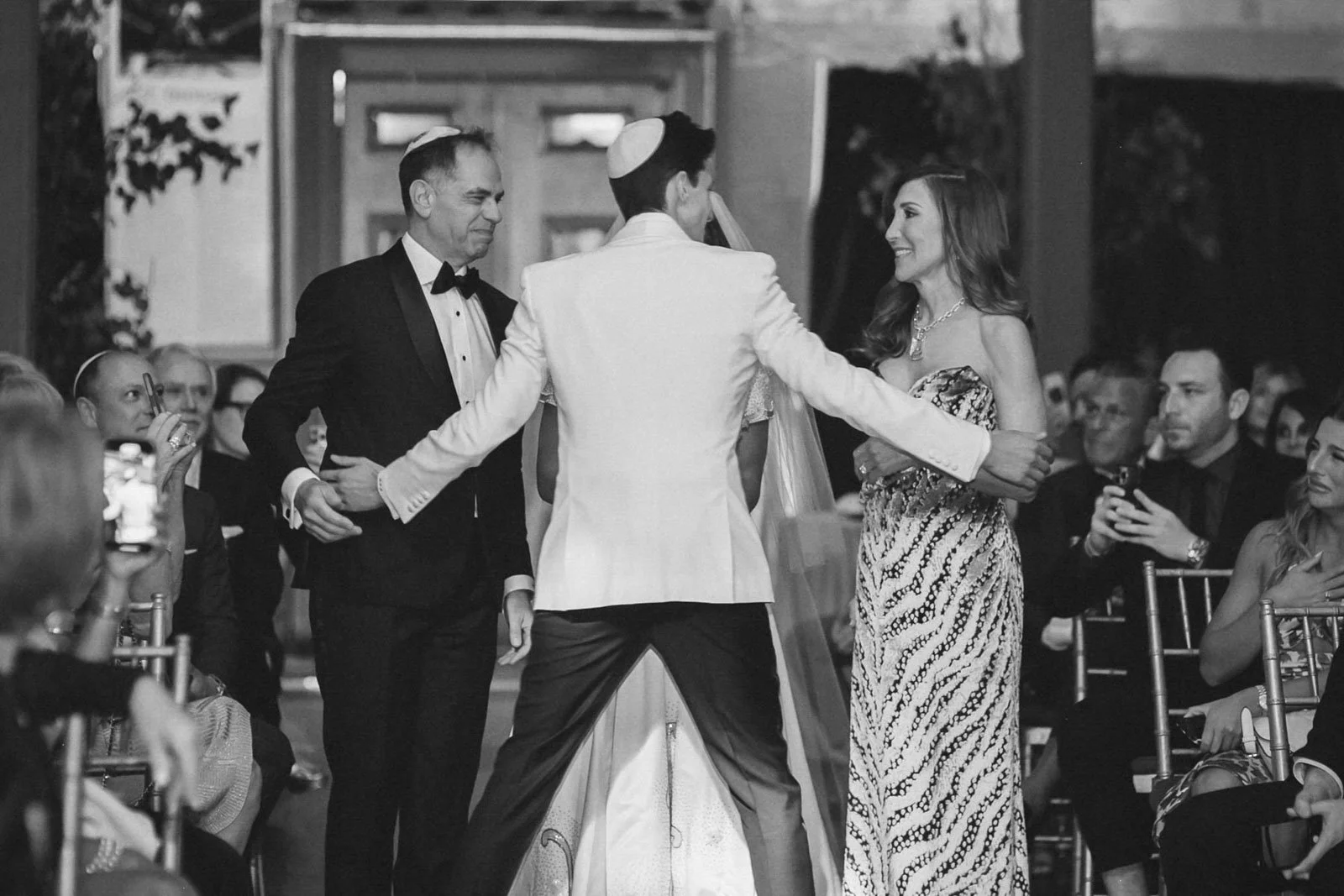 A black and white photo of a wedding ceremony with a groom in a white tuxedo and a bride in a patterned gown, standing and holding hands, facing each other, with guests watching and taking photos.