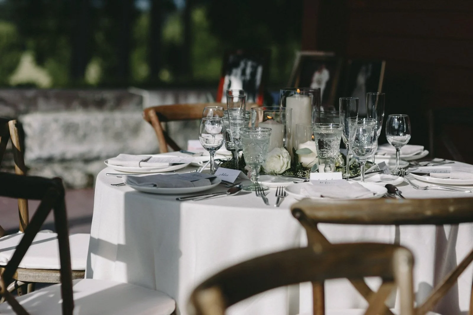 Elegant outdoor dining table set with white tablecloth, white floral centerpiece, candles, and glassware, surrounded by wooden chairs.