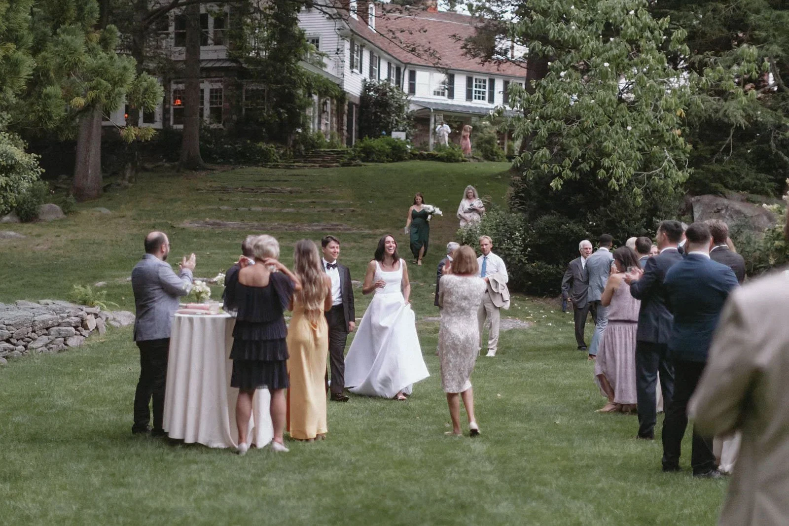 Guests celebrating a wedding outdoors on a lush green lawn in front of a large house, some clapping and smiling, with the bride in a white wedding gown and groom in a black tuxedo, surrounded by family and friends.