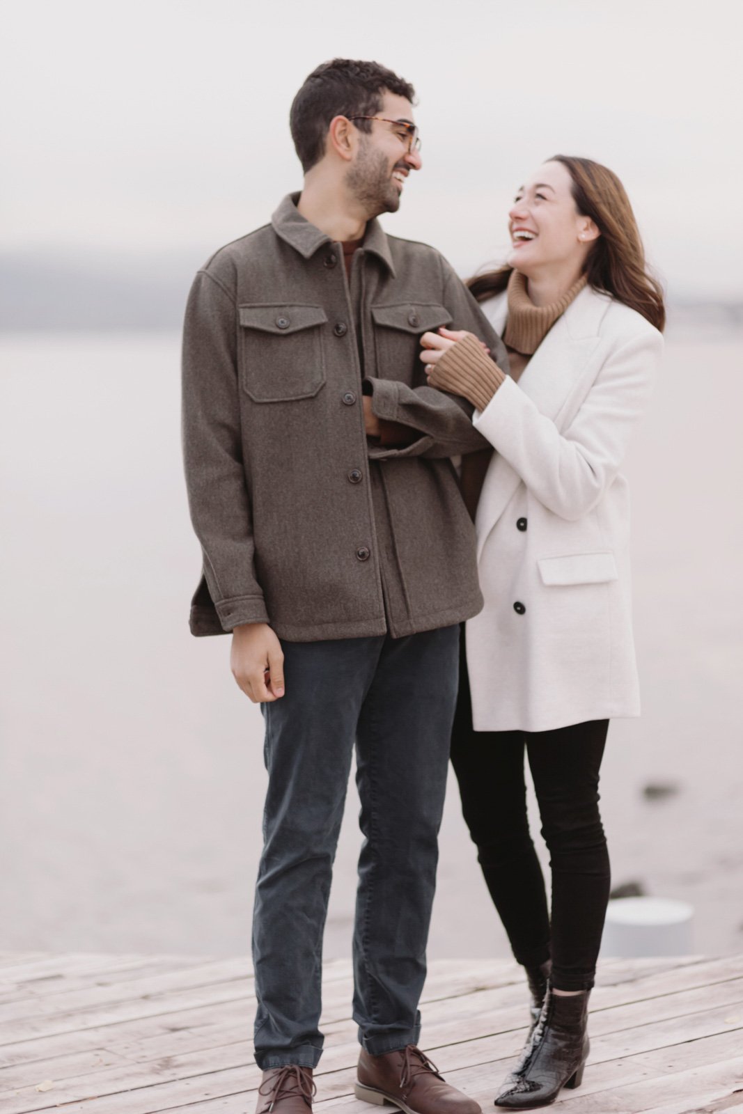 A man and woman smiling and laughing, standing on a wooden dock by a body of water, dressed in casual winter clothing.
