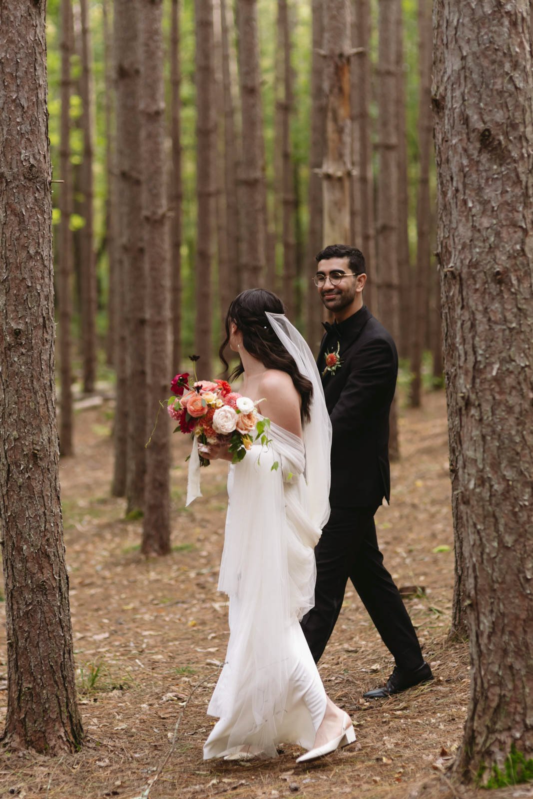 A bride and groom standing in a forest, with the bride holding a bouquet of flowers and wearing a white dress, and the groom in a black suit smiling at her.