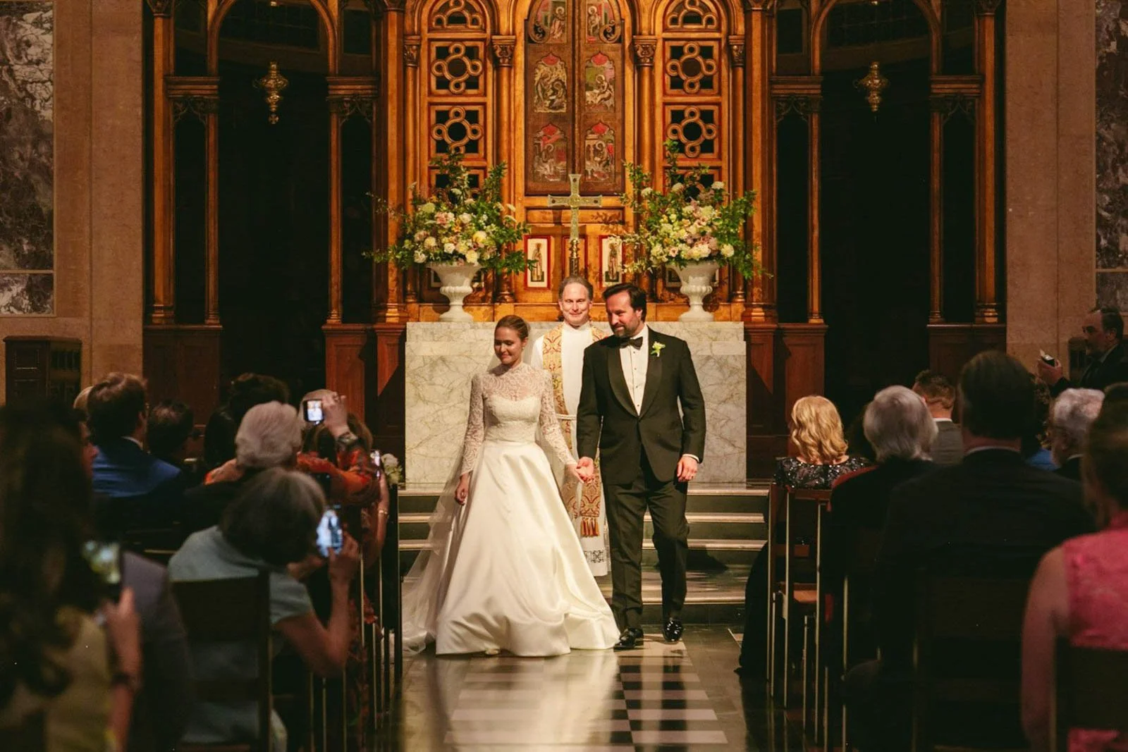 A bride and groom walk hand in hand down the aisle after a wedding ceremony in a church, with guests taking photos and watching.