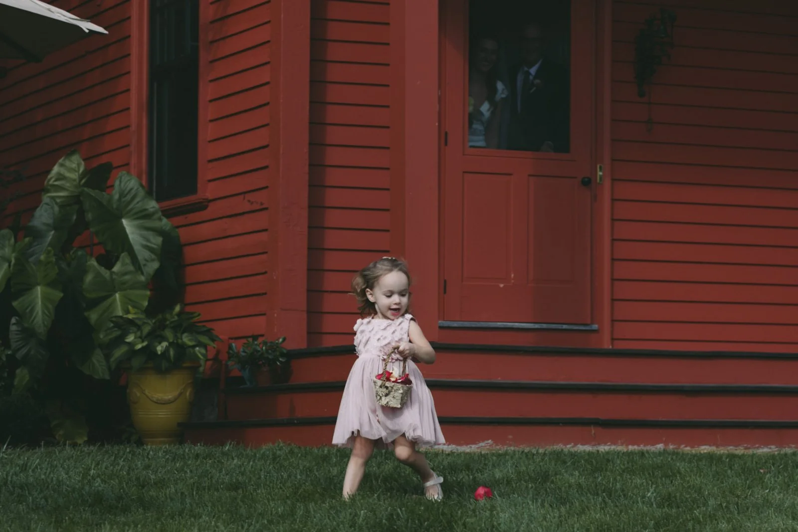 A young girl in a light pink dress playing outside on the grass with a small basket in her hand, near a red wooden house. Two people are visible inside the house through a window, looking out at the girl.