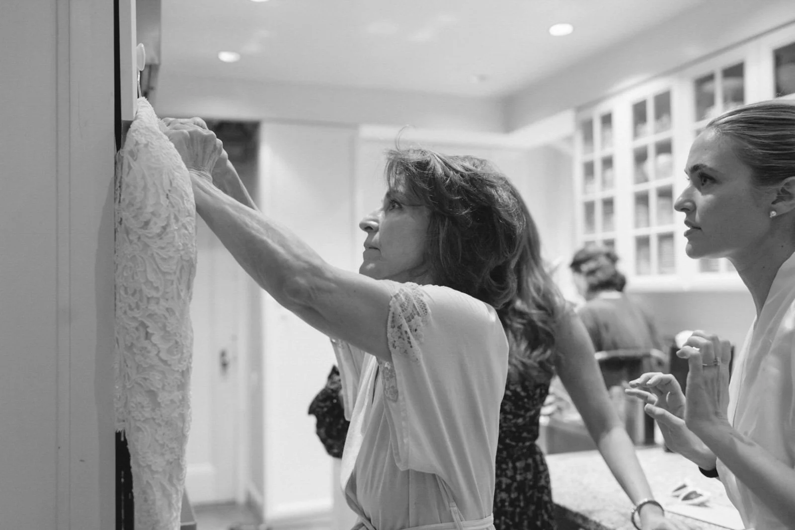 A woman with curly hair looks at a dress on a hanger while a blonde woman in a white shirt looks on in a room with bright lighting and other people in the background.