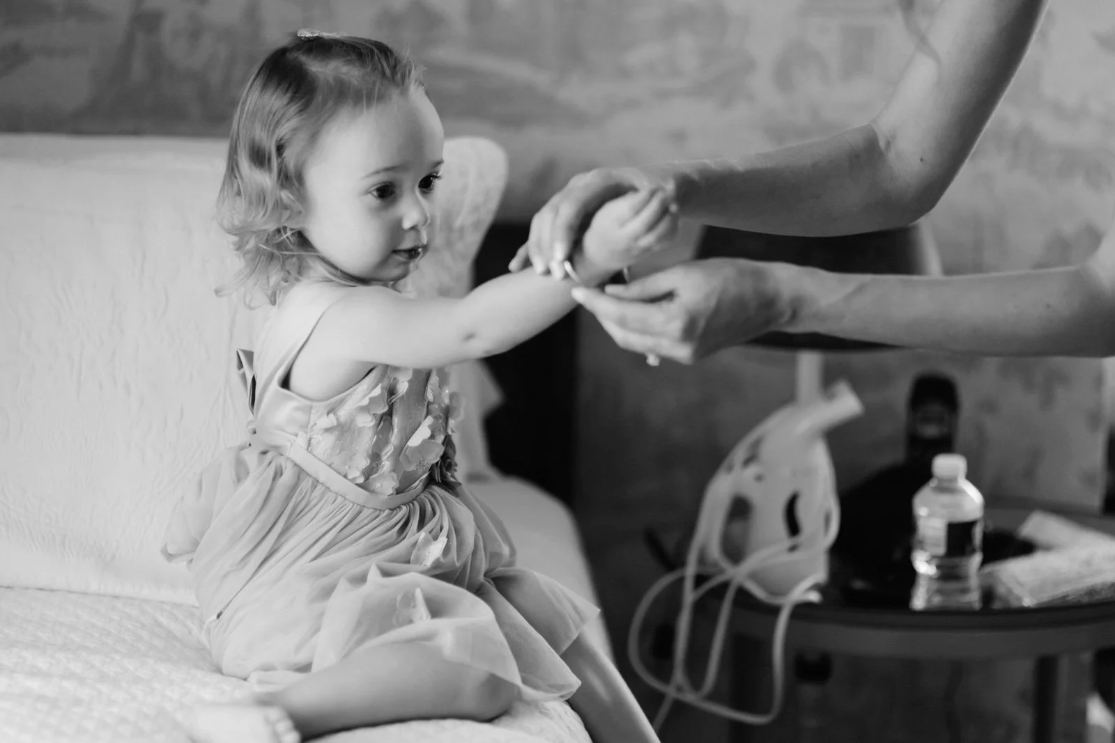 A young girl sitting on a bed, reaching out to hold an adult's hand in a black-and-white photo.