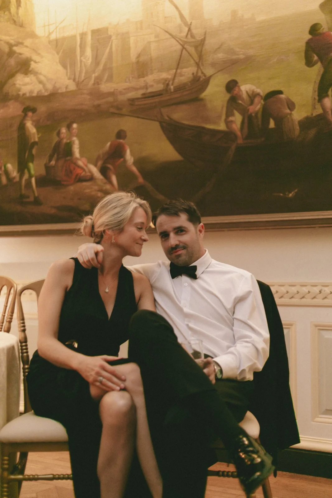 A couple sits close together at a formal event, with a woman leaning her head on the man's shoulder, both smiling. The man is wearing a white shirt and bowtie, the woman is in a black dress. Behind them is a large painting of people on a boat by a po