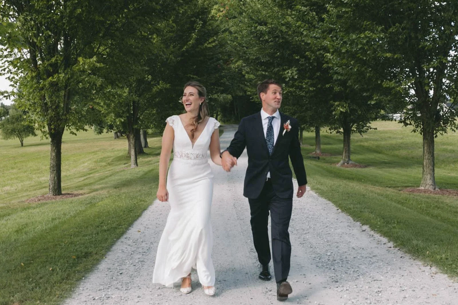 A happy bride and groom walking hand in hand on a gravel path through a park with green grass and trees.