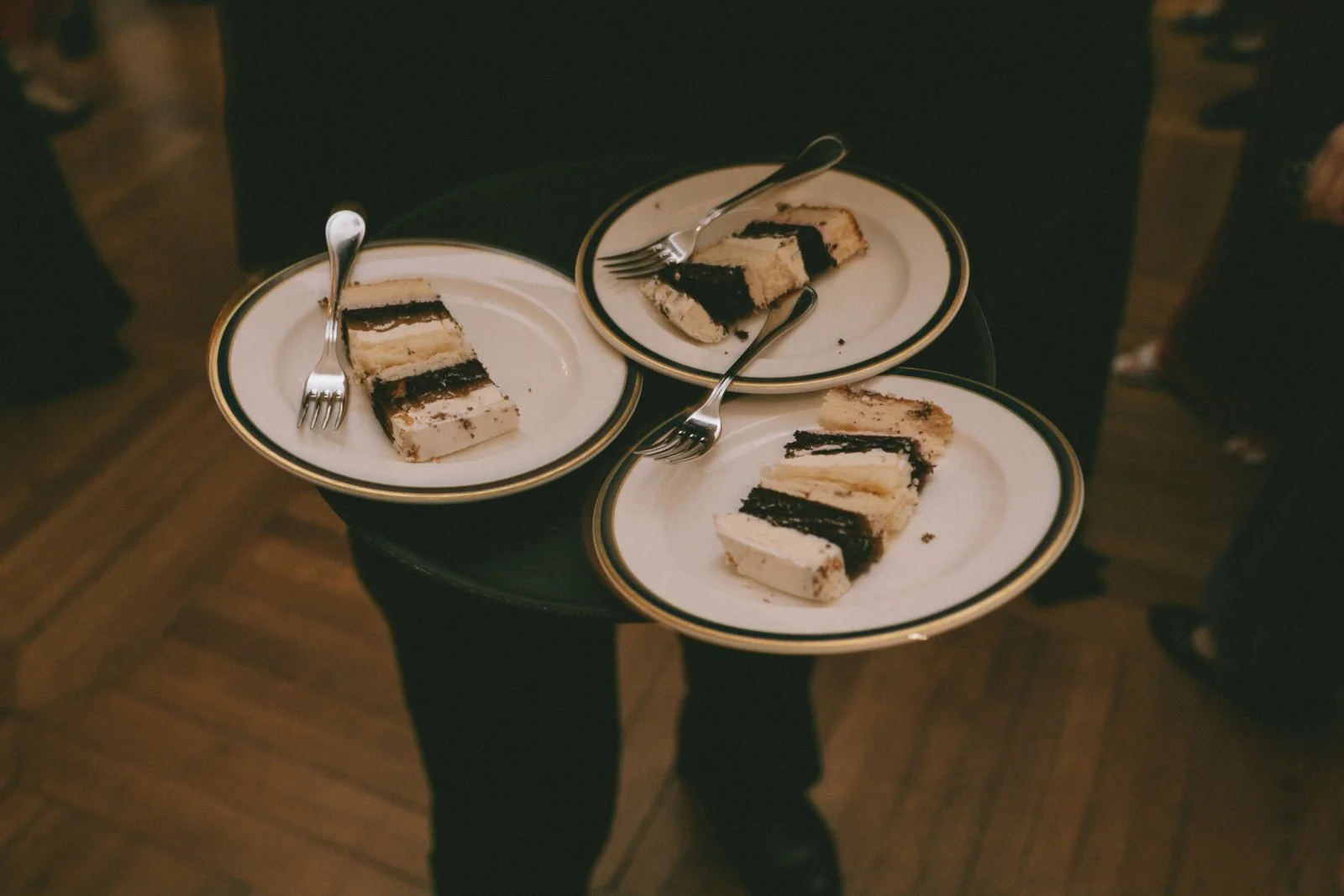 Three white plates with slices of black and white layered cake, each with a fork, held by a person in dark clothing over a wooden floor.