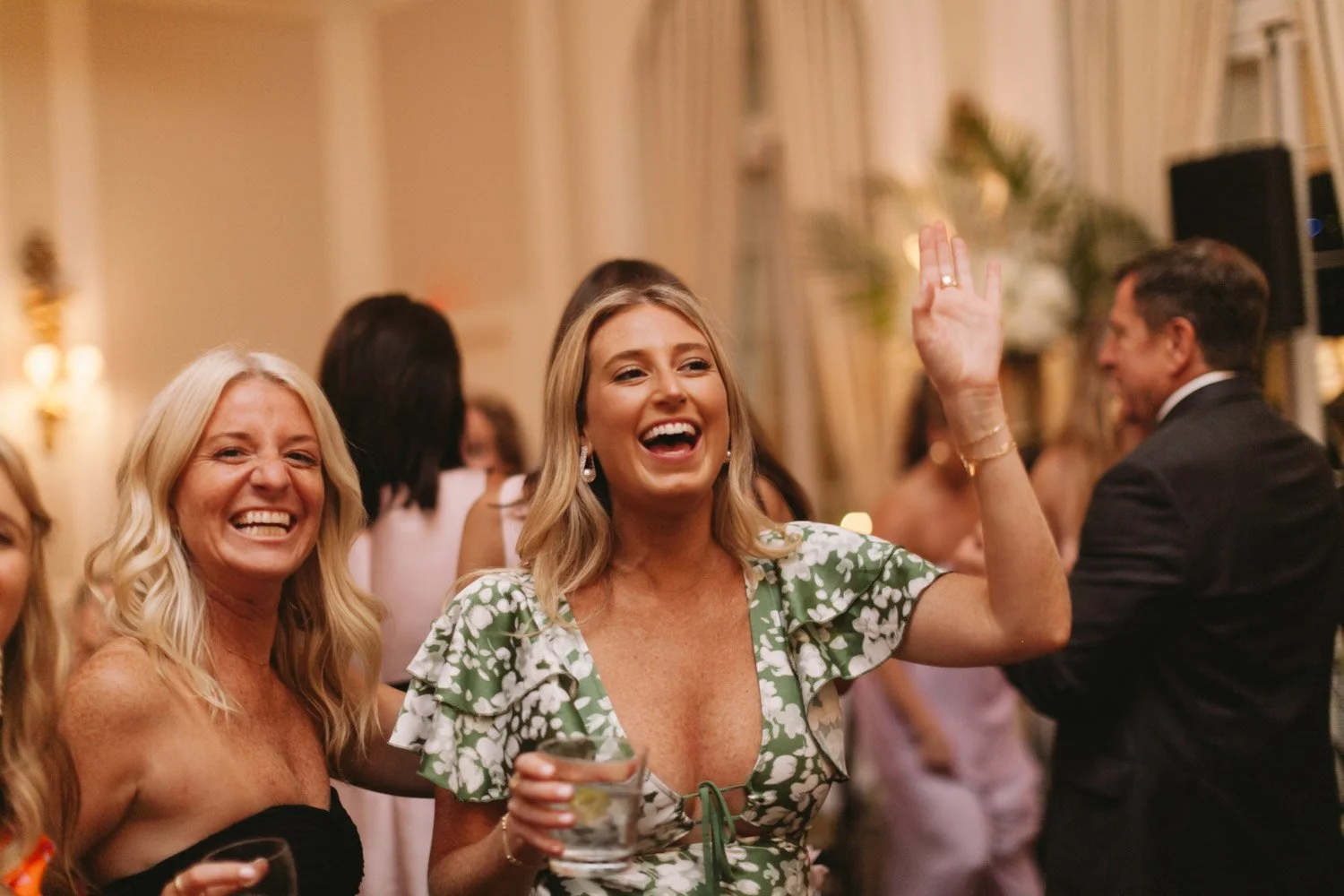 People celebrating at a party, including a woman in a floral dress waving and holding a drink, and another woman smiling along with others in the background.