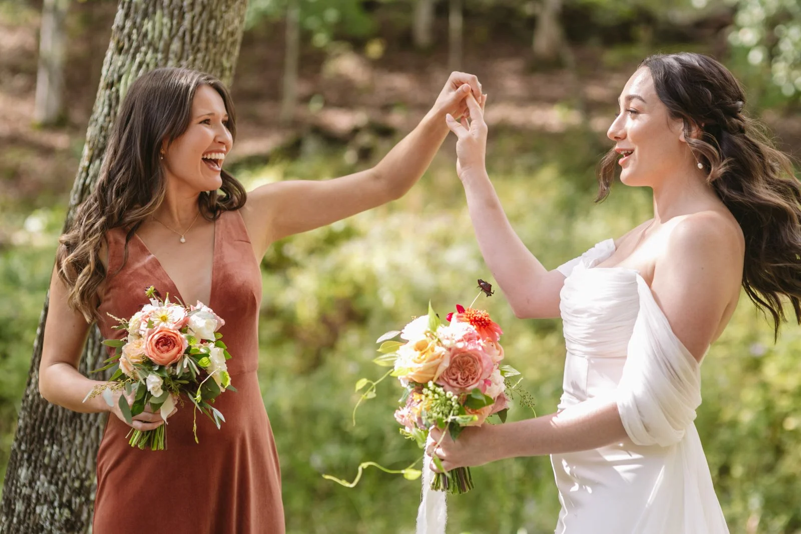 Two women in a forest, one in a white wedding dress and the other in a rust-colored dress, holding bouquets of flowers, smiling, and touching hands.