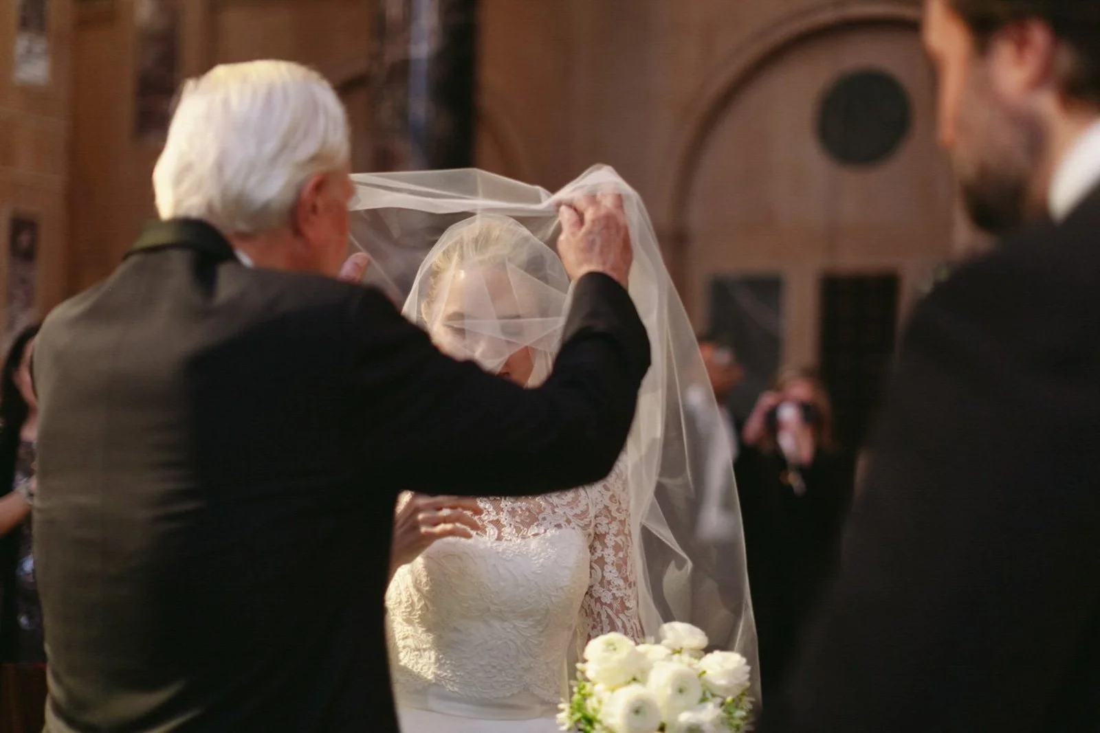An elderly man helps a bride with her veil during a wedding ceremony, with a bouquet of white flowers on the table nearby.
