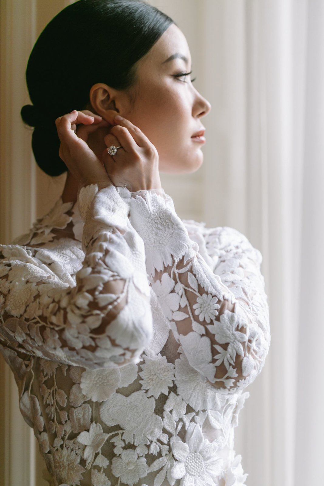 Woman with dark hair styled in a low bun, wearing a white, floral, lace dress, putting on earrings.