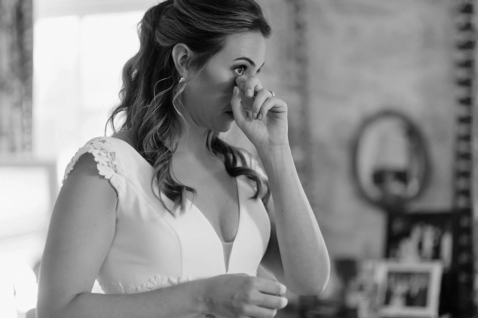 A woman with wavy hair in emotional distress, holding her nose with her hand, in a room with a mirror and photo frames in the background.