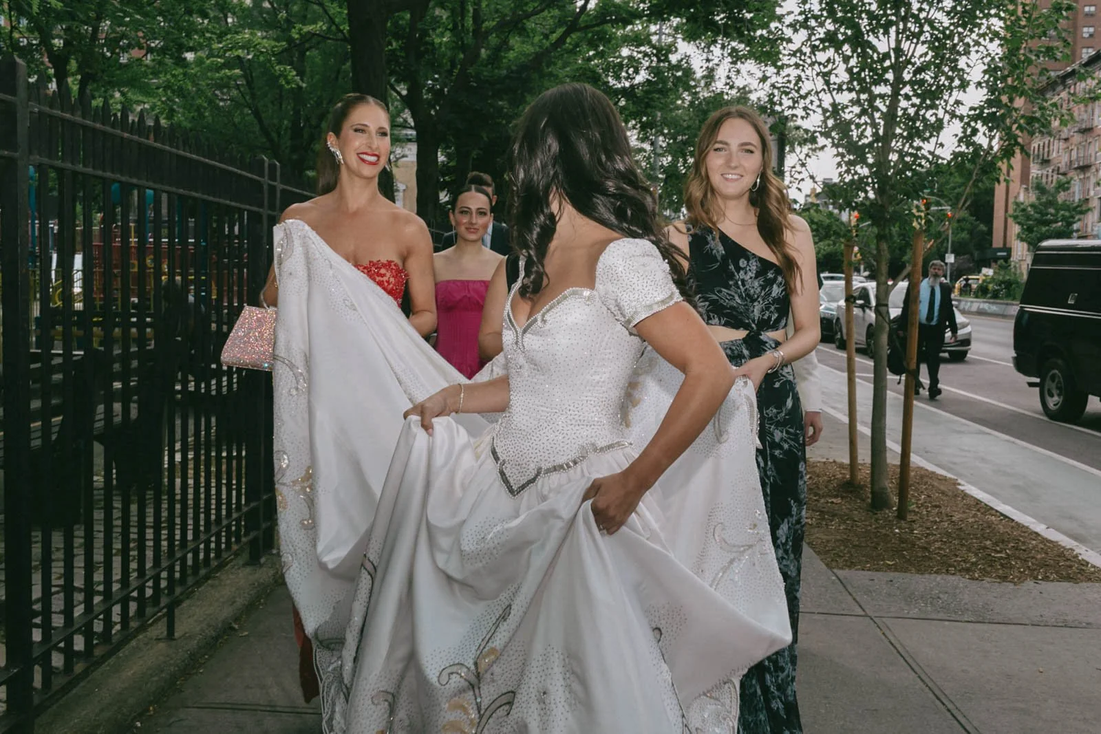 Group of women dressed in formal and colorful attire standing on sidewalk near fence with trees and cars in the background