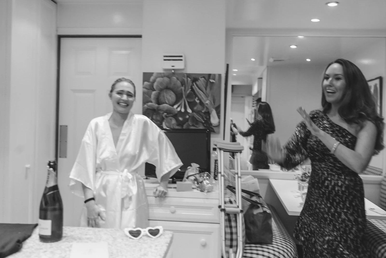 Two women smiling and laughing in a room, one wearing a satin robe and the other in a sparkly dress, with a woman in the background taking a photo.