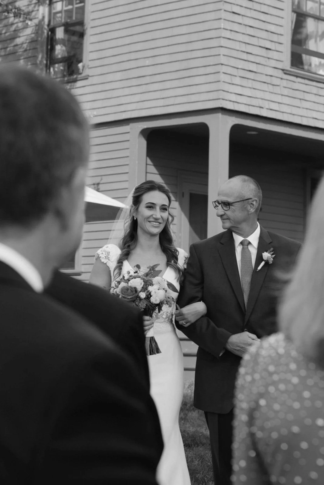 A bride holding a bouquet of flowers smiling as she walks with an older man in a suit, with other wedding guests partially visible in the foreground, outside a house with siding and windows.