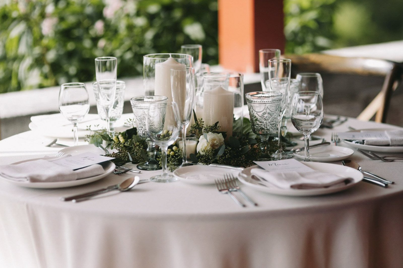 Elegant outdoor dining table set with white plates, silverware, wine glasses, water glasses, candles, and floral decor.