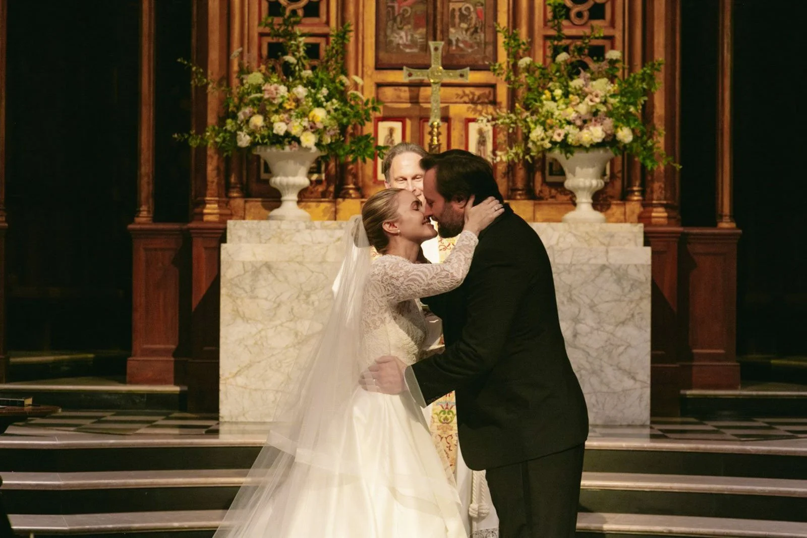 A bride and groom sharing a kiss during their wedding ceremony in a church, with a priest behind them and floral arrangements on the altar.