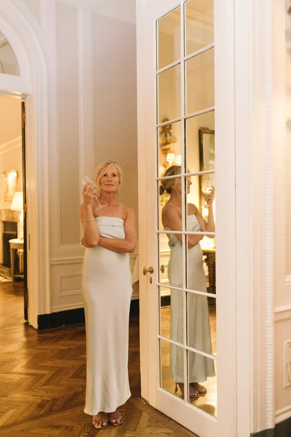 A woman in a white strapless dress holding a wine glass stands next to a large window with multiple panes, with her reflection visible in the window. The setting appears to be a well-lit, elegant room with wooden flooring and classic decor.