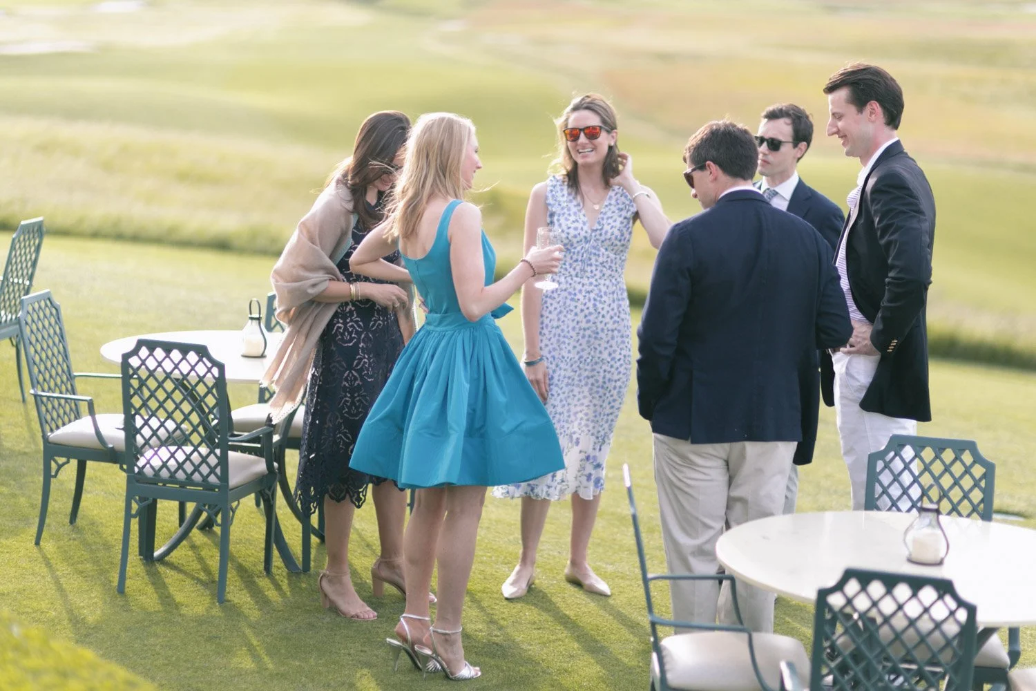 Group of six well-dressed people socializing outdoors on a sunny day, standing on a grassy lawn near tables and chairs.