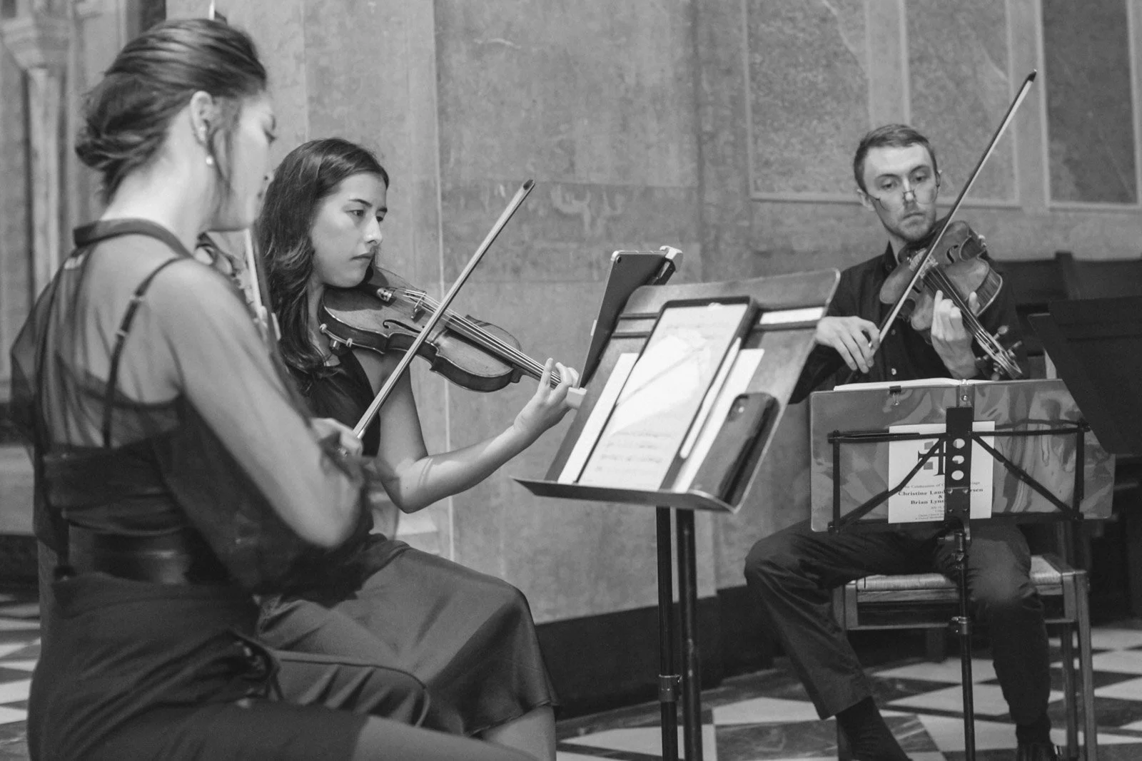 Three musicians playing violins during a performance, two women and one man, seated in a room with stone walls and checkered flooring.