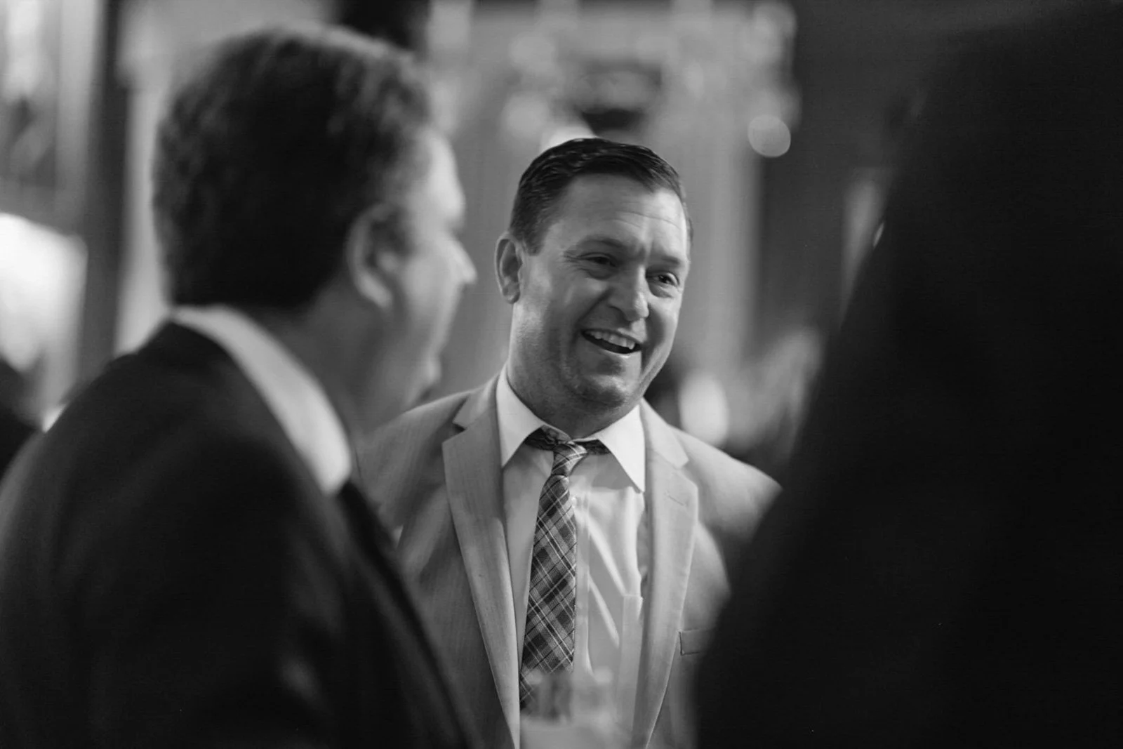Black and white photo of three men in suits smiling and conversing at a social gathering.