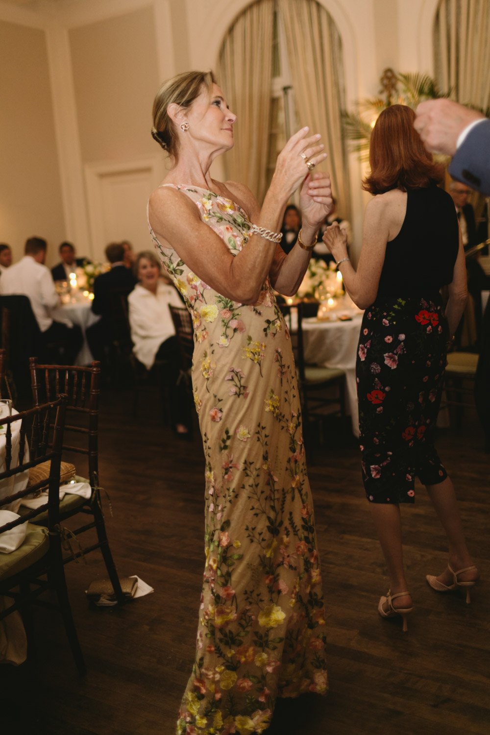 A woman dancing at a formal event or celebration, wearing a floral dress and jewelry, with people seated at tables and a decorated dining area in the background.